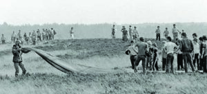A crowd gathers in a field to watch a paratrooper fold his parachute just after he was airdropped from a U.S. Air Force C-130 during exercise Reforger/Autumn Forge 1980.  Autumn Forge is a reserve airdrop taking place here and in Germany and involving mostly U.S. and British troops and equipment.
